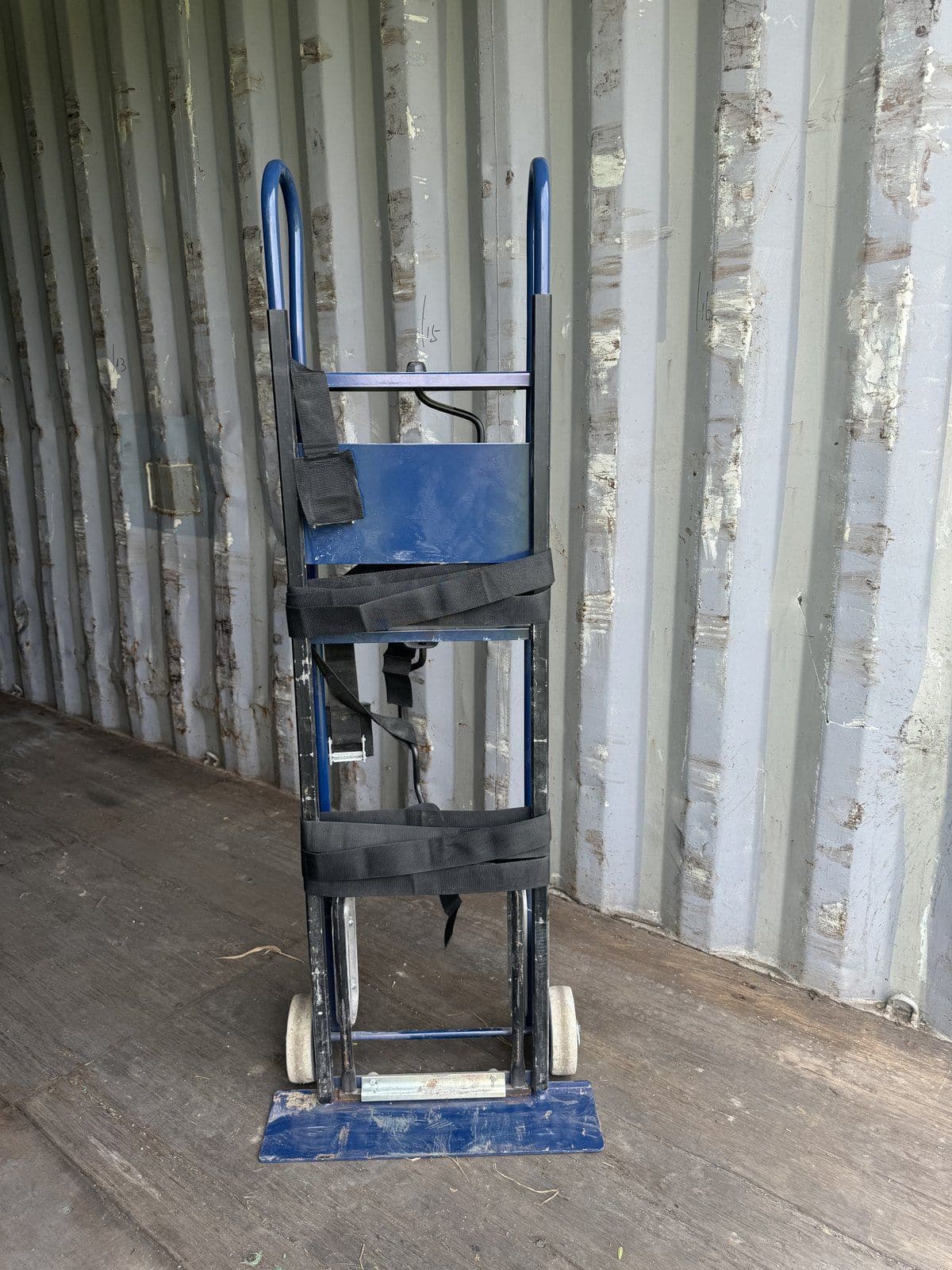 Blue appliance hand truck with black securing straps, standing in a storage container