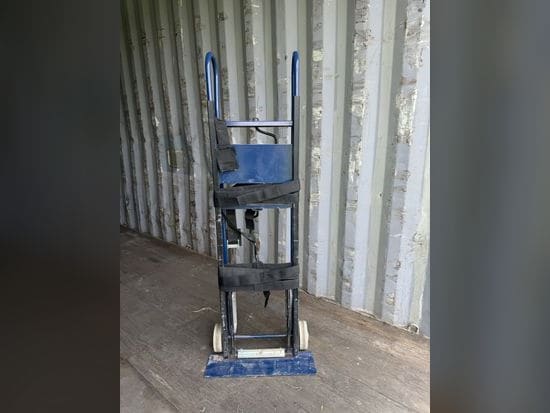 Blue appliance hand truck with black securing straps, standing in a storage container