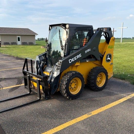 John Deere 318G skid steer with pallet forks, parked on pavement